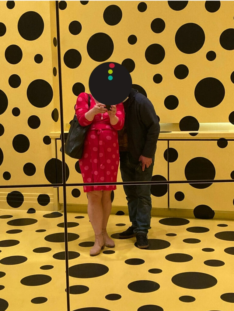 Zwei Personen vor gelber Wand mit schwarzen Punkten, eine trägt rotes Kleid mit weißen Punkten, die andere dunkle Kleidung, beide spiegeln sich in einer Spiegelfläche. Die Köpfe der Personen sind von einem schwarzen Punkt verdeckt