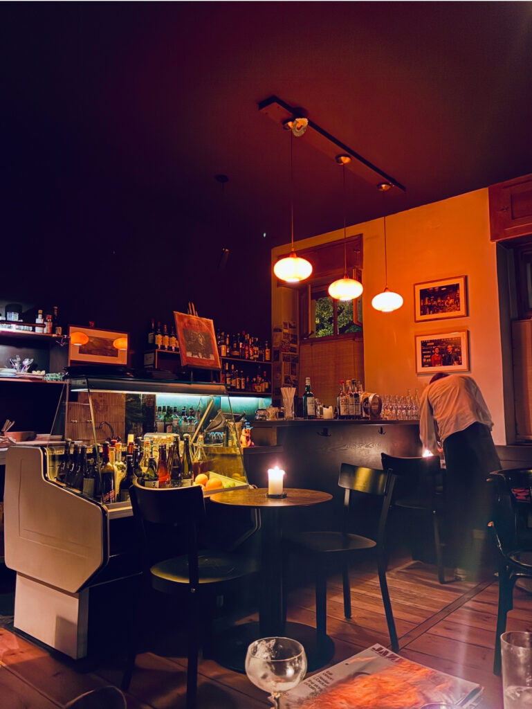 Gemütliche Bar mit runden Tischen, Stühlen, einer brennenden Kerze auf einem Tisch und einer Person, die an der Theke arbeitet