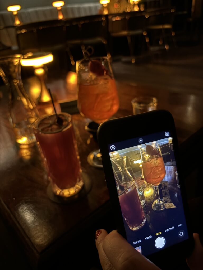 Hand hält Smartphone, das zwei Getränke auf einem dunklen Tisch in einer Bar zeigt, auch die Getränke selbst sind zu sehen