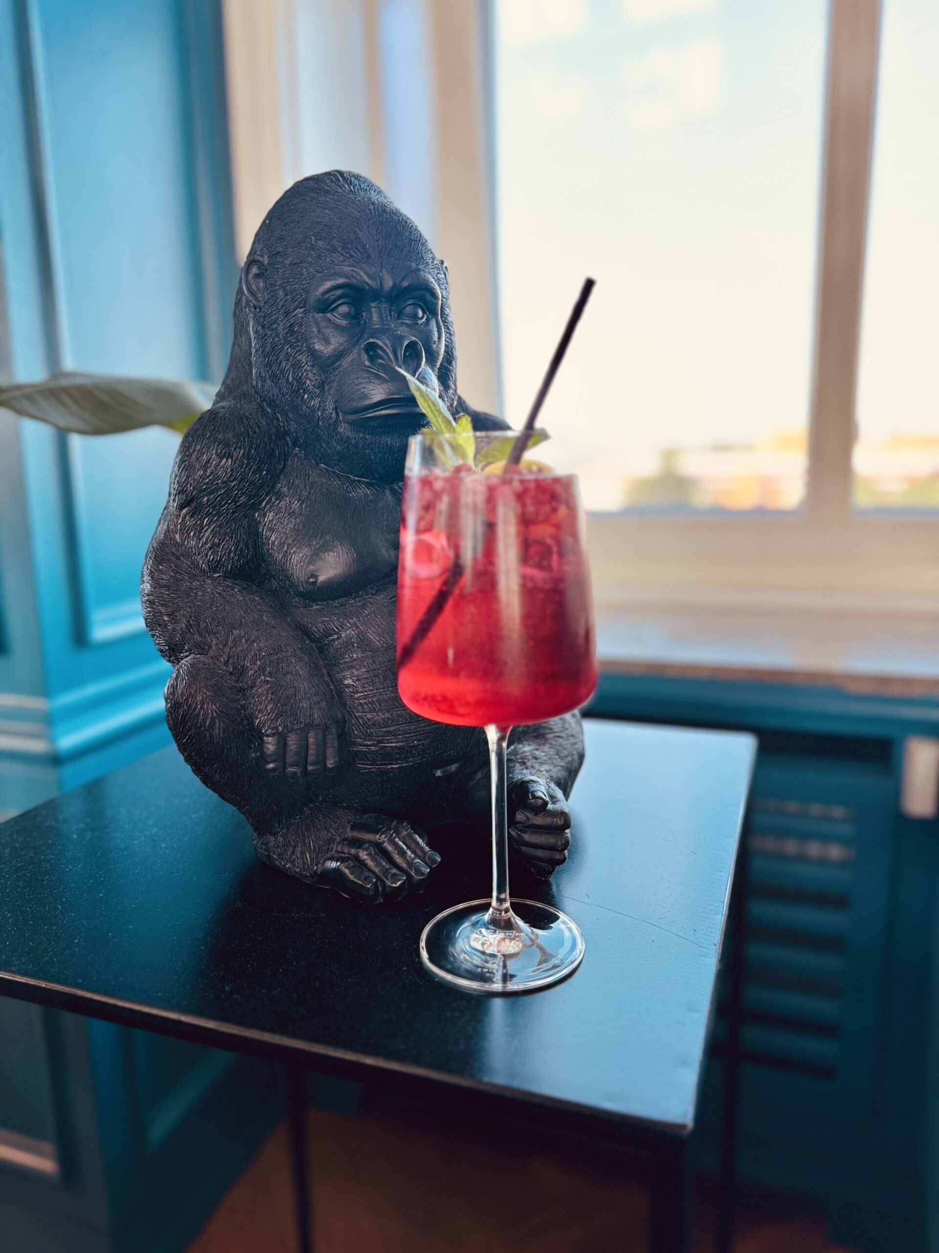 Schwarze Gorillafigur sitzt auf einem Tisch neben einem Glas mit rotem Getränk und Strohhalm