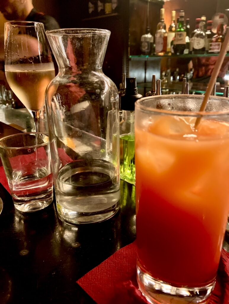 Glas mit orangerotem Cocktail auf rotem Untersetzer neben leerer Karaffe und Sektglas auf dunklem Tresen in Bar mit Spirituosen im Hintergrund