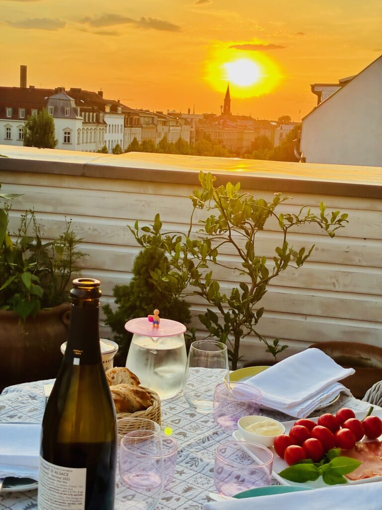 Sonnenuntergang über einer Stadt mit gedecktem Tisch auf einer Terrasse, darauf Weinflasche, Gläser, Brot, Tomaten und Servietten