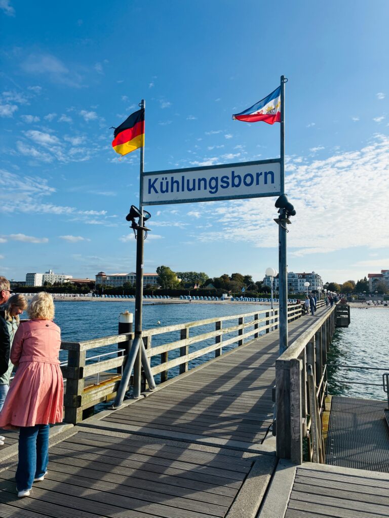Steg mit Schild 'Kühlungsborn' und deutschen sowie polnischen Flaggen über Wasser mit Menschen am linken Rand