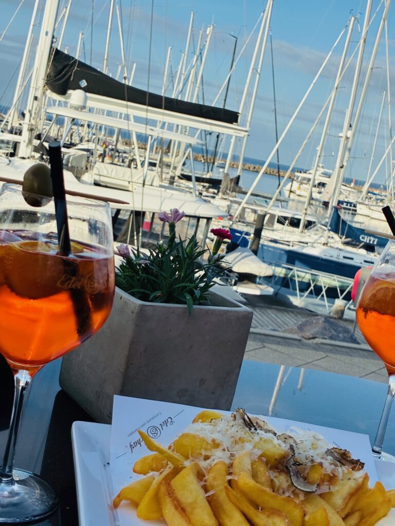 Zwei Gläser mit orangem Getränk, ein Teller mit Pommes und Trüffeln, ein Blumentopf und im Hintergrund Segelboote im Hafen
