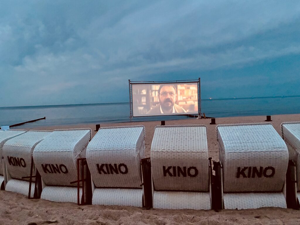 Strand mit mehreren weißen Strandkörben mit der Aufschrift 'KINO' vor einer Leinwand, auf der ein Mann mit Brille und Bart zu sehen ist