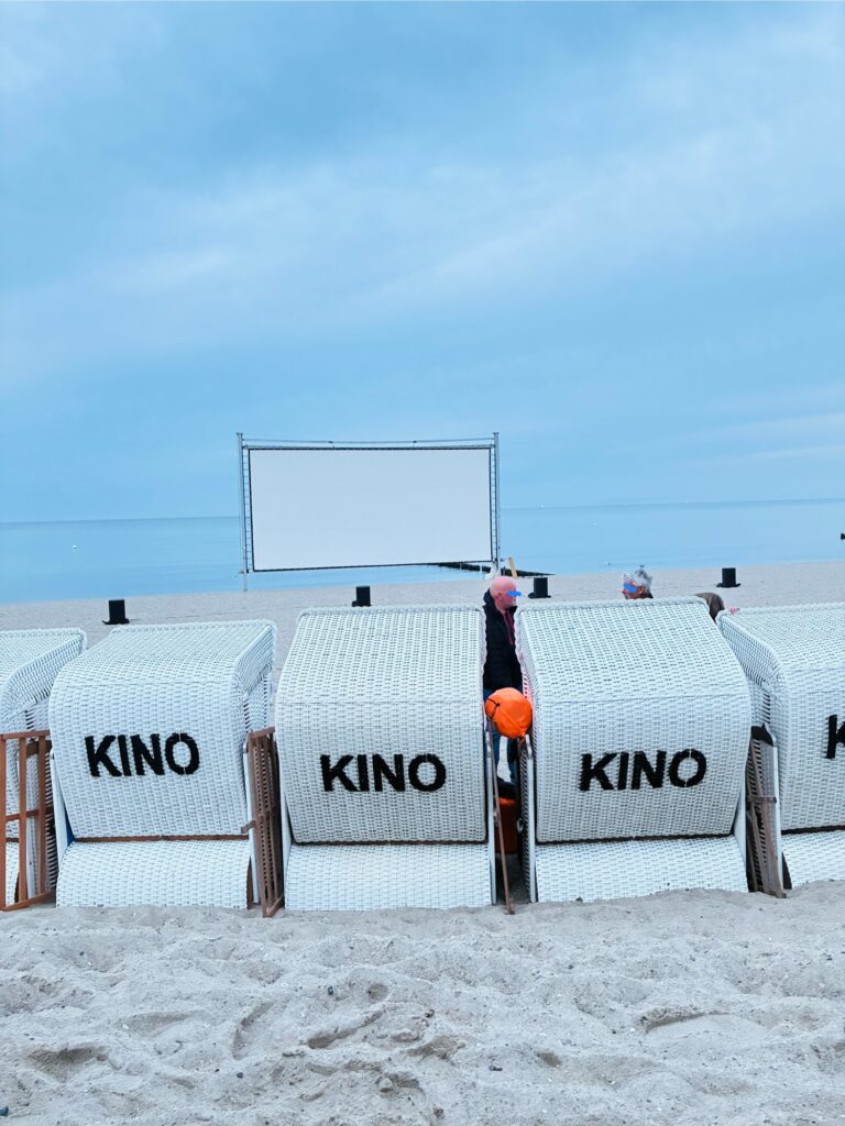 Mehrere weiße Strandkörbe mit der Aufschrift 'KINO' stehen am Sandstrand vor einer großen weißen Leinwand