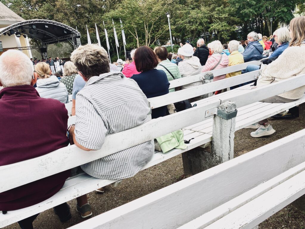 Menschen sitzen auf weißen Holzbänken im Freien vor einer Bühne mit schwarzem Bühnenüberdachung und weißen Fahnen im Hintergrund