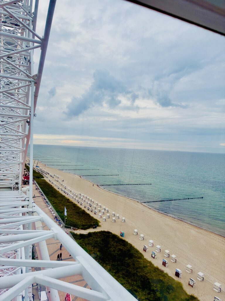 Blick durch das Gestänge eines Riesenrades auf einen Strand mit Reihen von Strandkörben und das Meer unter bewölktem Himmel