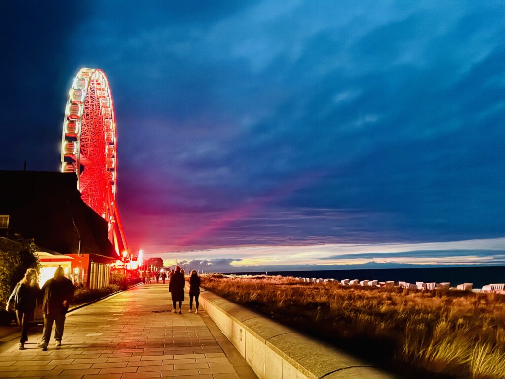Menschen gehen am Abend auf einer Promenade mit beleuchtetem Riesenrad links und Strandkörben rechts unter bewölktem Himmel