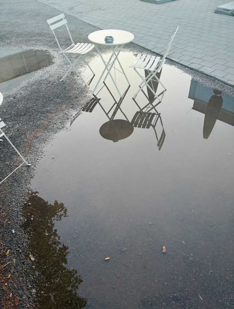 Zwei weiße Stühle und ein runder Tisch mit Aschenbecher spiegeln sich in einer großen Pfütze auf Kies