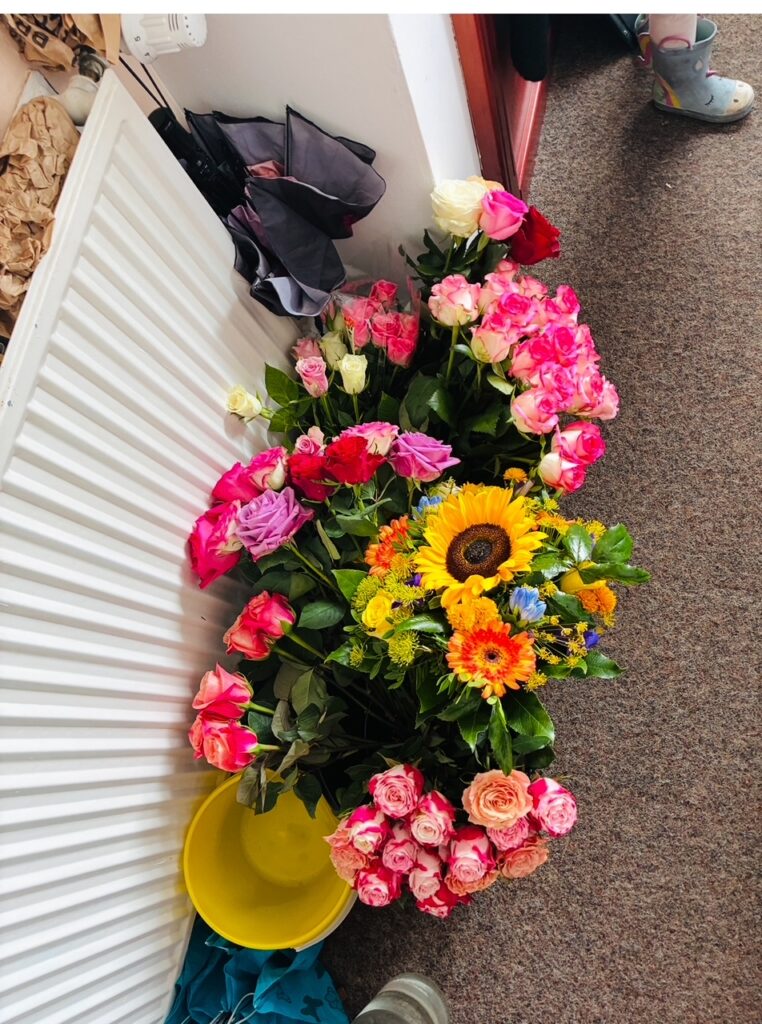 Bunte Blumensträuße mit Rosen und Sonnenblumen stehen auf dem Boden neben einem Heizkörper und einem gelben Eimer