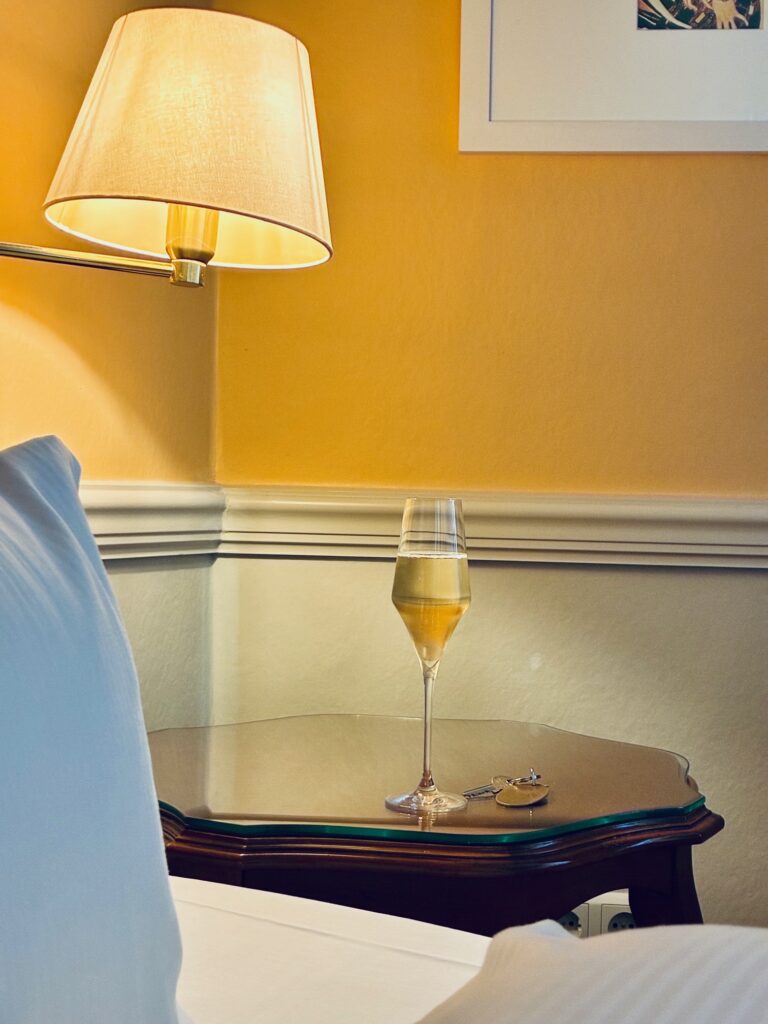 Volles Sektglas auf einem dunklen Nachttisch neben einem weißen Bett und einer Wandlampe an gelber Wand