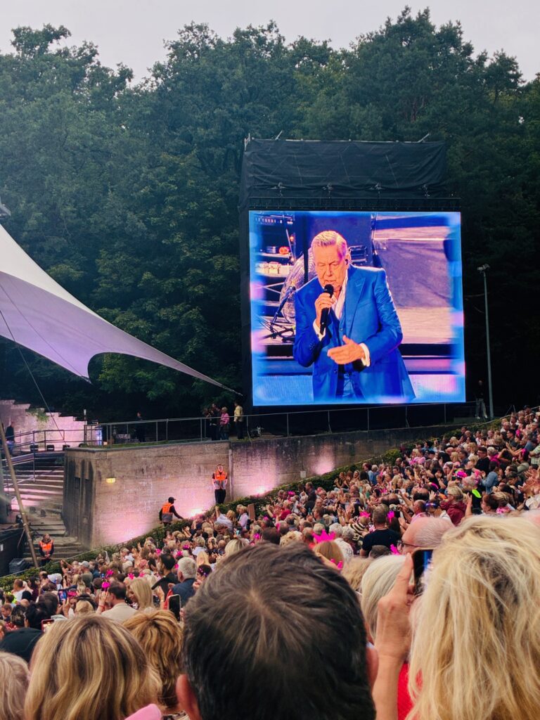 Große LED-Leinwand zeigt Sänger in blauem Anzug vor Publikum vor Freiluftbühne mit Bäumen im Hintergrund. Der Sänger ist Roland Kaiser.