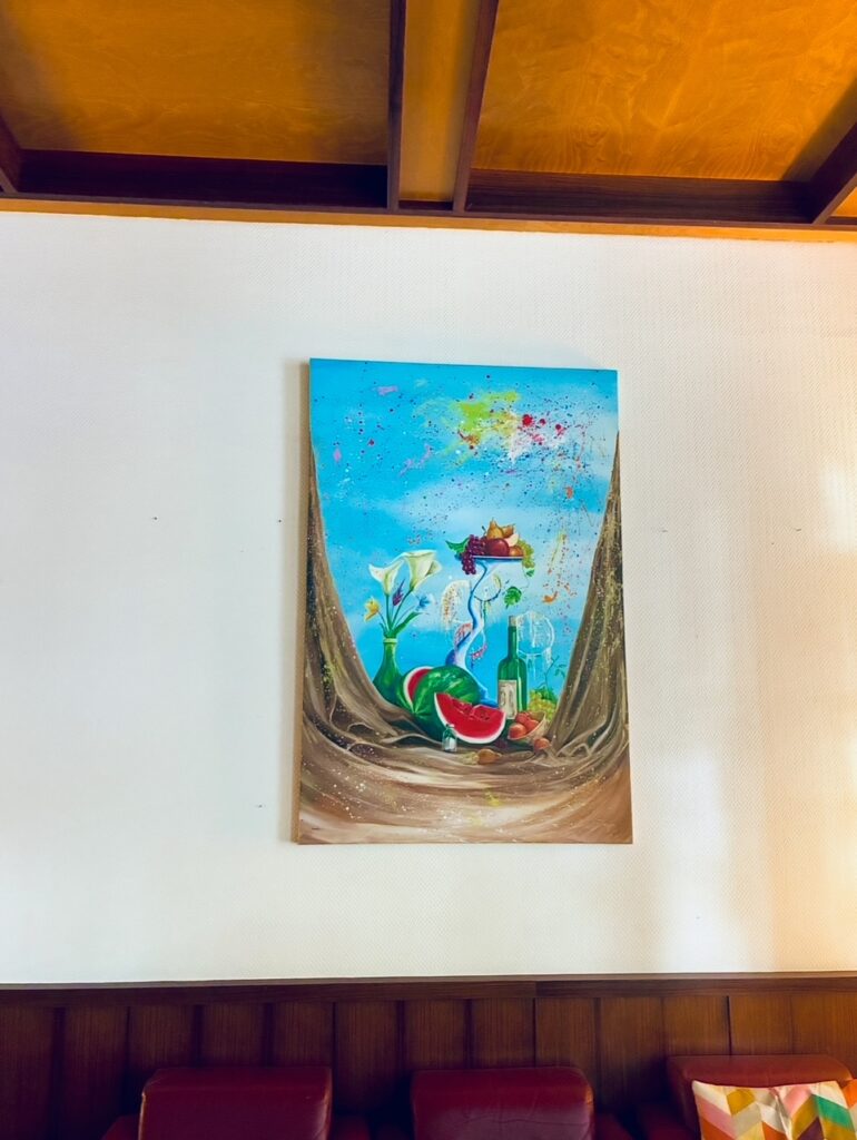 Gemälde an weißer Wand mit blauem Himmel, Blumen, Wassermelone, Flasche und Früchten, darüber Holzbalken und darunter braune Holzvertäfelung mit Sitzbank
