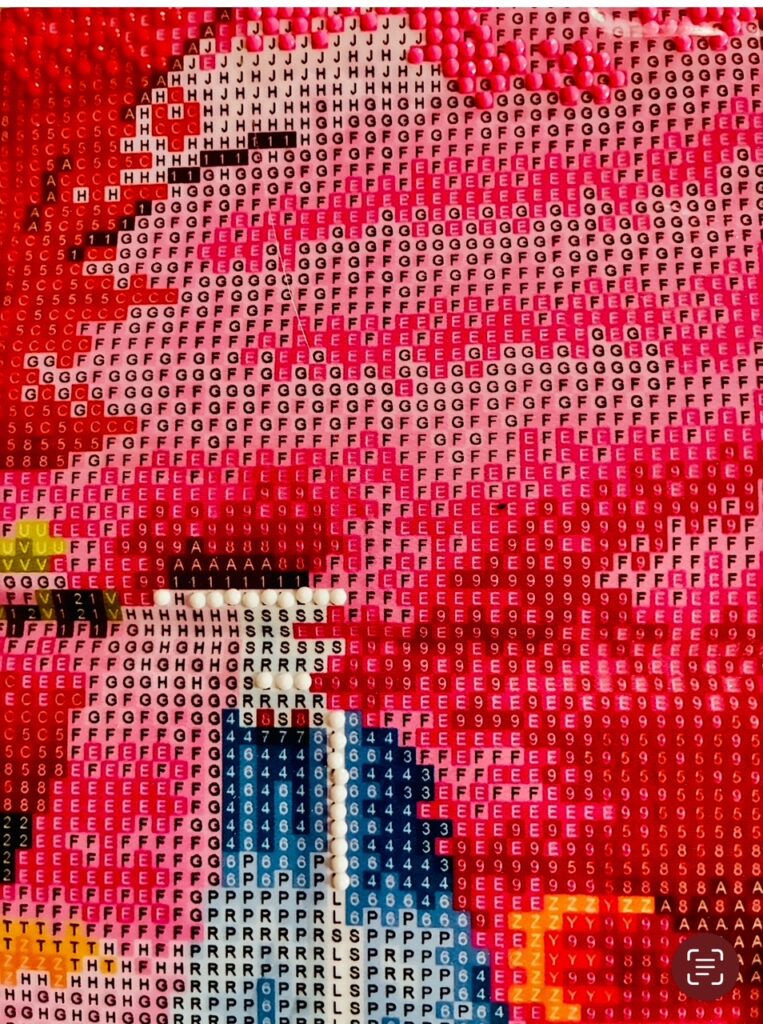 Nahaufnahme einer pixelartigen Darstellung mit Buchstaben und Zahlen in verschiedenen Farben, es handelt sich um eine unfertige Diamond Painting-Arbeit