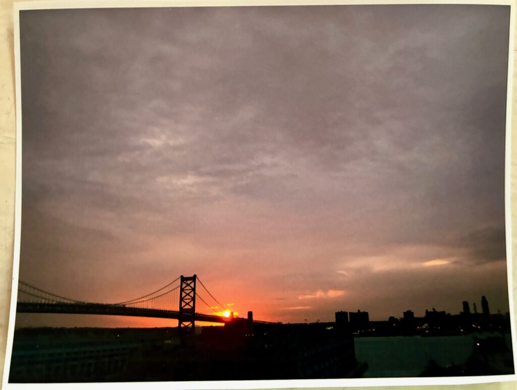 Abbild einer analogen Fotografie eines Sonnenuntergangs hinter einer Hängebrücke mit dunkler Stadtsilhouette und bewölktem Himmel