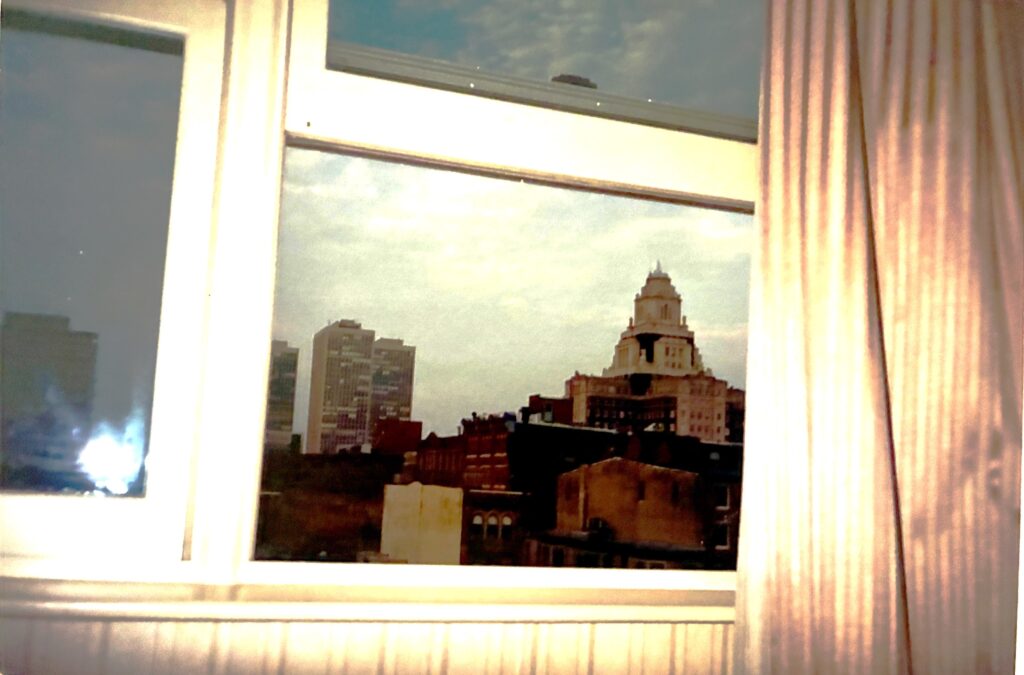Analoge Fotografie einer Stadtansicht mit mehreren Hochhäusern durch ein Fenster mit weißen Rahmen und halb geöffneten weißen Vorhängen