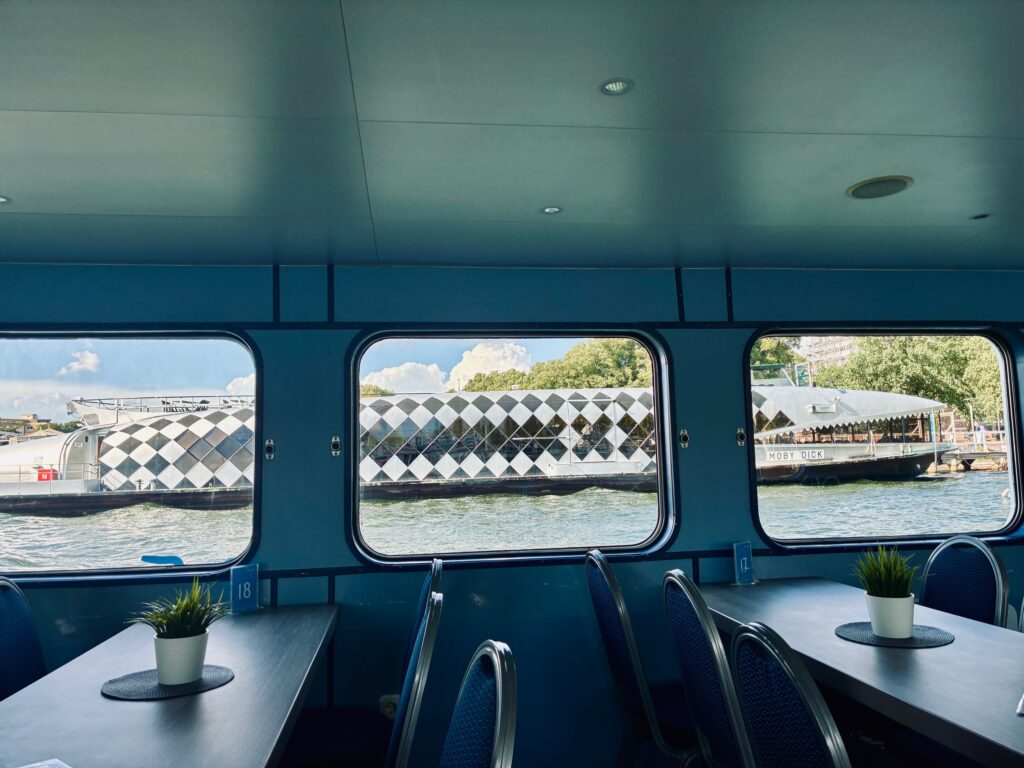 Blick aus dem Inneren eines Schiffes mit blau gepolsterten Stühlen und einem Tisch mit kleinem Pflanzengefäß auf das Wasser und ein weiß-schwarz gemustertes Ausflugsboot in Form eines Wals außerhalb der Fenster
