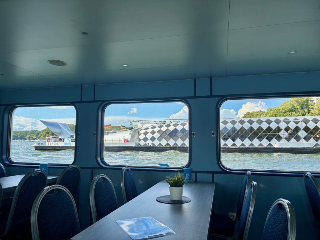 Blick aus dem Inneren eines Schiffes mit blau gepolsterten Stühlen und einem Tisch mit kleinem Pflanzengefäß auf das Wasser und ein weiß-schwarz gemustertes Ausflugsboot in Form eines Wals außerhalb der Fenster
