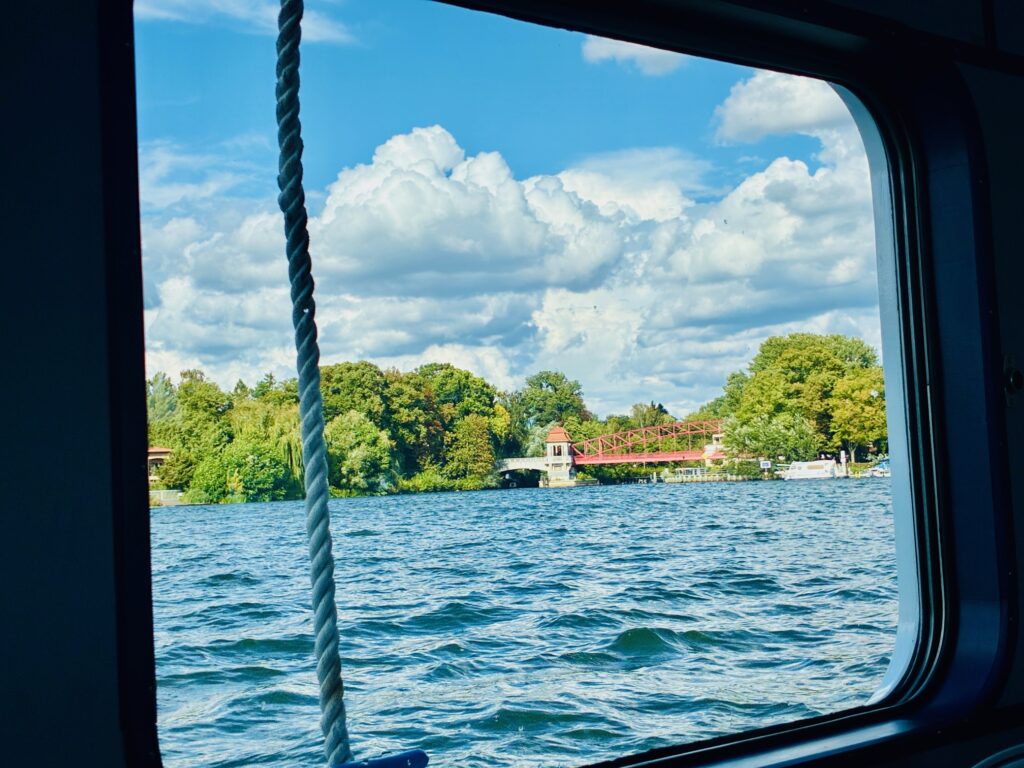 Blick durch ein rechteckiges Bootsfenster auf einen See mit bewaldetem Ufer und einer roten Brücke im Hintergrund