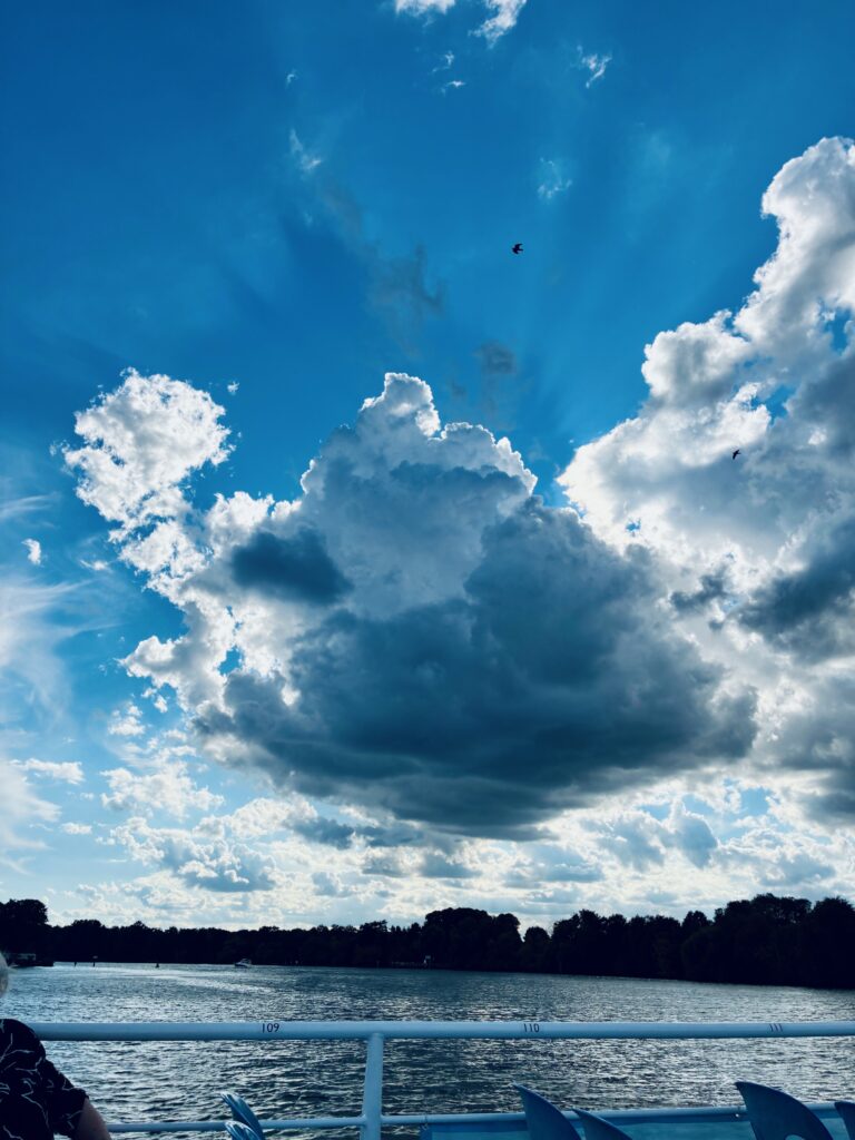 Wolken am blauen Himmel über einem See mit Baumreihe am Ufer und Teil eines Boots mit Geländer und Stühlen im Vordergrund