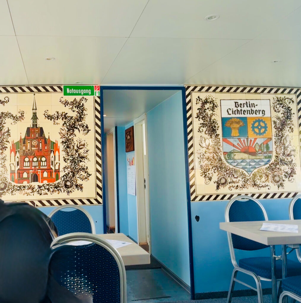 Innenraum eines Ausflugsschiffes mit blauen Stühlen und Tischen vor zwei Wandbildern, links ein historisches Gebäude, rechts das Wappen von Berlin-Lichtenberg, darüber ein grünes Notausgangsschild