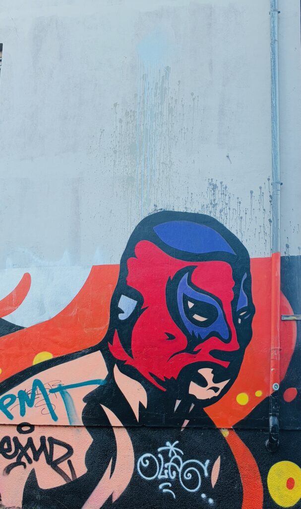 Wandgraffiti des stilisierten Gesichts eines Superhelden mit Wrestlermaske mit roter und blauer Farbgebung und schwarzen Konturen vor orangefarbenem Hintergrund