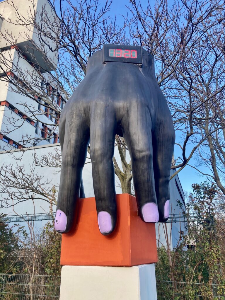 Große schwarze Hand mit lila Fingernägeln auf einem weißen und orangen Sockel vor kahlen Bäumen und einem brutalistischen Schulgebäude. Es handelt sich um die Skultpur "Hand mit Uhr" von Joachim Schmettau